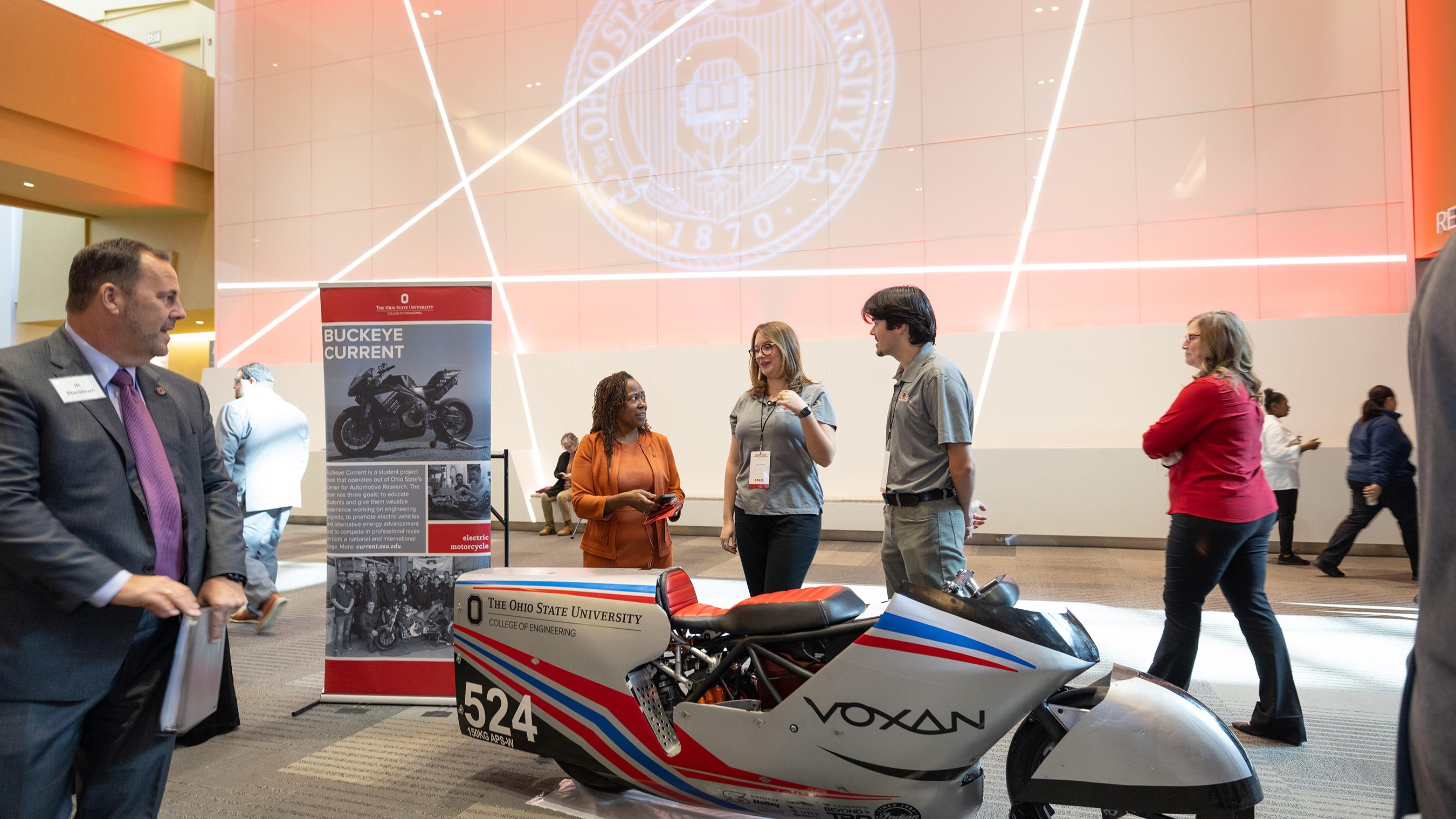 People talking about the electric vehicles in front of projected university seal