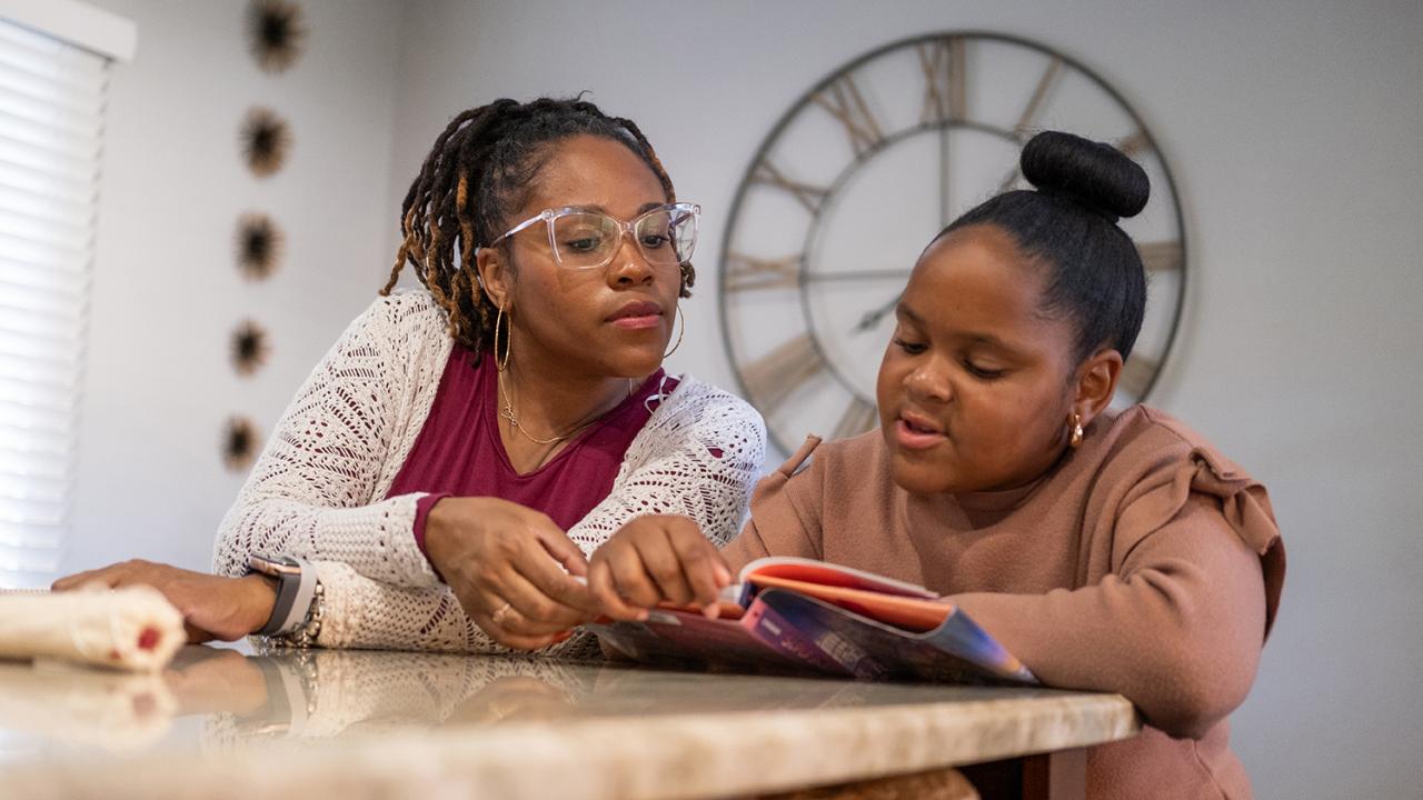 A mother and her daughter read a book together at a table.